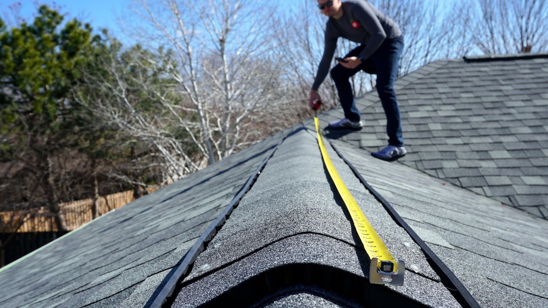 Roofer measuring roof ridge with yellow tape measure on shingle roof